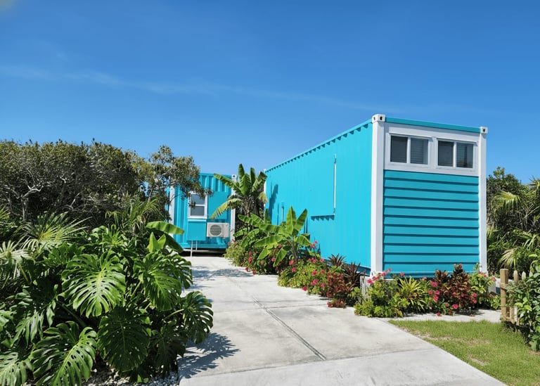 Bright turquoise and white modern container home with tropical landscaping and palm trees under clear blue sky